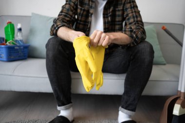 Adam oturma odasında kanepede otururken temizlik için sarı lastik eldiven giyiyor. Ev işleri. Yakın plan.