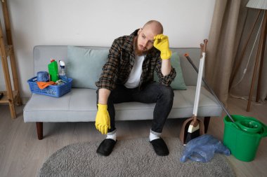 Eldivenli yorgun adam kanepeye oturuyor ve evi temizledikten sonra dinleniyor. Boşluğu kopyala