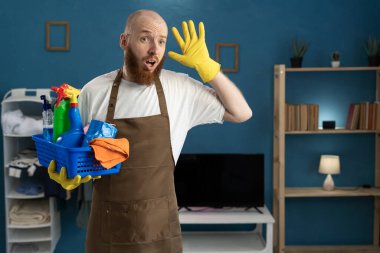 Şaşırmış sakallı, negatif duyguları olan temizlikçi adam oturma odasında temizlik sepetiyle dikiliyor. temizlik hizmeti ve ev işi konsepti