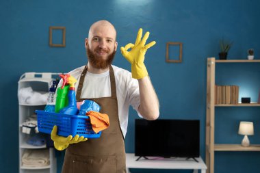 Önlük ve eldiven giyen mutlu, pozitif sakallı adam renkli deterjan şişeleriyle dolu bir jest sepeti gösteriyor. Oda servisi ve ev işi kavramı