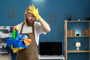 Üzüntülü erkek temizlikçi kafasında hoşnutsuz bir ifadeyle temizlik malzemeleriyle dolu bir sepet tutuyor. Temizlik, angarya ve ev işleri.