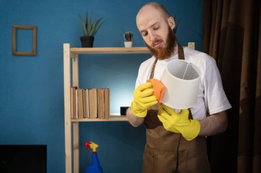 Sakallı adam oturma odasındaki toz beziyle lambaları yıkıyor ve temizliyor. Boşluğu kopyala