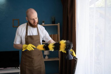 Evdeki oturma odasını temizlerken önlüklü bir adam tüy fırçasıyla toz alıyor. Günlük temizlik, temizlik, yüzey bakımı ve uygun aletlerle ev bakımını gösteriyor.