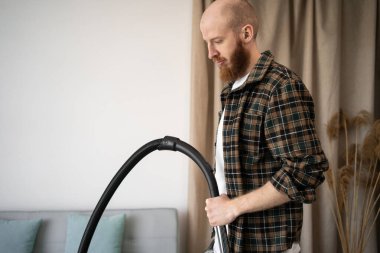 Evdeki oturma odasında ekose gömlek giyen sakallı adam. Temizlikçi evdeki temizlik işleri için yerleri süpürüyor. Boşluğu kopyala