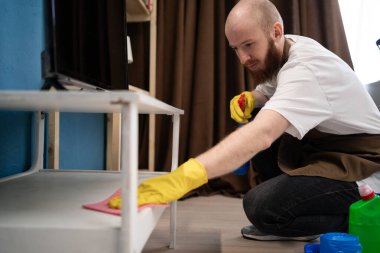 Adam evdeki oturma odasındaki paçavrayla raftan tozu kaldırıyor. Erkek hademe mobilyaları mikrofiber bez ile siliyor. Temizlik ve angarya konsepti