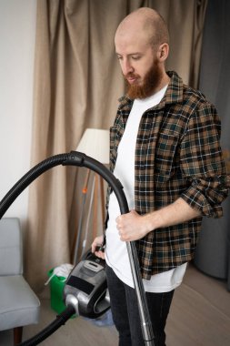 Evdeki oturma odasında ekose gömlek giyen sakallı adam. Temizlikçi evdeki temizlik işleri için yerleri süpürüyor. Boşluğu kopyala