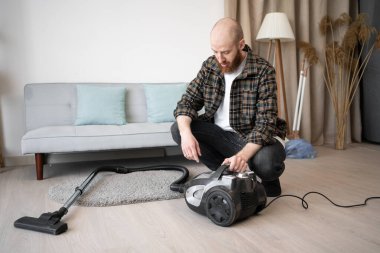 Sakallı adam oturma odasında elektrikli süpürgeyle halıyı süpürmeyi bitiriyor. Ev işleri, ev işleri ve ev işleri konsepti. Boşluğu kopyala
