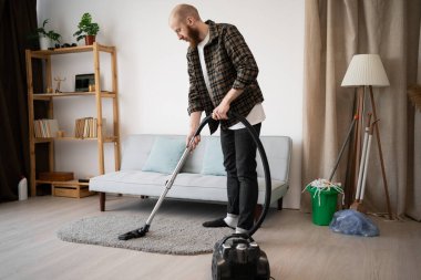 Adam oturma odasında elektrikli süpürgeyle halı süpürüyor. Ev sahibi elektrikli ev aletlerini kullanarak ev işi yapıyor. Boşluğu kopyala