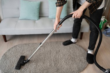Adam oturma odasında elektrikli süpürgeyle halı süpürüyor. Ev sahibi elektrikli ev aletlerini kullanarak ev işi yapıyor. Yakın plan.