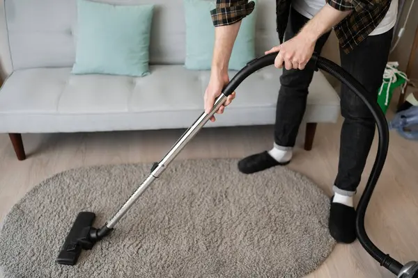 Adam oturma odasında elektrikli süpürgeyle halı süpürüyor. Ev sahibi elektrikli ev aletlerini kullanarak ev işi yapıyor. Yakın plan.