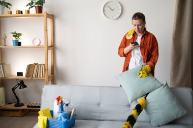 Temizlik yaptıktan ve akıllı telefon kullandıktan sonra kanepeye uzanan kadın modern teknoloji, rahatlama, dijital hayat ve ev bakımı kavramının karışımını gösteriyor.