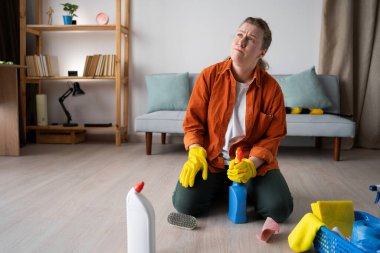 Temizlik malzemeleriyle yerde oturan genç bir kadın üzgün ve mesafeli görünüyor, ev temizliği ve günlük ev işleri sırasında stres, depresyon ve duygusal mücadele belirtileri gösteriyor. Boşluğu kopyala
