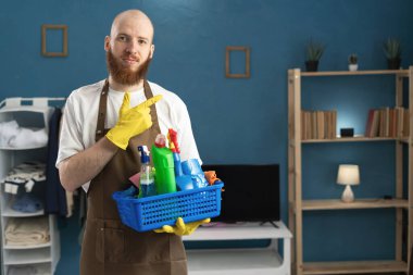 Eldiven ve önlük takan genç sakallı bir adam temizlik malzemeleriyle mesaj için yeri işaret ediyor. Ev işlerinde erkek hizmetçi. Boşluğu kopyala