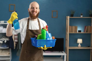 Gülümseyen sakallı bir adam eldiven ve önlük takıyor ve temizlik malzemeleriyle birlikte deterjan püskürtüyor. Ev işlerinde erkek hizmetçi. Ev temizliği, ev temizliği ve ev işi konsepti