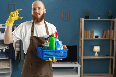 Eldiven ve önlük giyen sakallı genç bir adam. Temizlik malzemeleriyle birlikte. Parmaklarıyla küçük boy jest yapıyor. Ev işlerinde erkek hizmetçi. Ev temizliği, ev temizliği ve ev işi konsepti