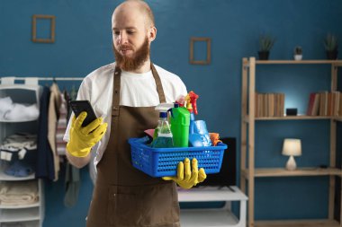 Eldiven ve önlük giyen sakallı genç adam. Yanında temizlik malzemeleriyle birlikte akıllı telefondan sosyal medya okuyor. Ev işlerinde erkek hizmetçi. Ev temizliği, ev temizliği ve ev işi konsepti
