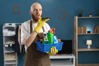 Eldiven takan sakallı genç adam ve temizlik malzemeleriyle dolu önlük sepetini tutan telefon kullanarak sesli mesajı dikte ediyor. Ev işlerinde erkek hizmetçi. Ev temizliği, ev temizliği ve ev işi konsepti