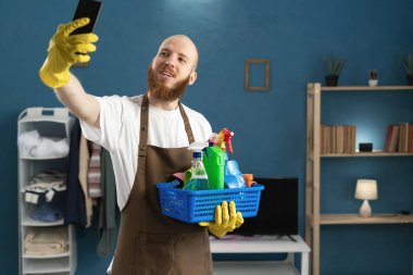 Eldiven ve önlük giyen, temizlik malzemeleriyle dolu sepet taşıyan genç sakallı adam kameraya kendi portresini çekiyor. Ev işlerinde erkek hizmetçi. Ev temizliği, ev temizliği ve ev işi konsepti