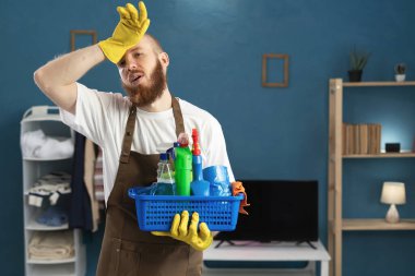 Eldiven giyen, eldiven takan, önlük tutan, temizlik malzemesi taşıyan yorgun bir adam. Stres ve depresyondan kafası ağrıyor. Ev işlerinde erkek hizmetçi. Boşluğu kopyala