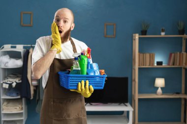 Şok olmuş, eldivenli ve önlüklü bir adam elinde temizlik malzemeleriyle zaferi kutluyor. Ev işlerinde erkek hizmetçi. Boşluğu kopyala