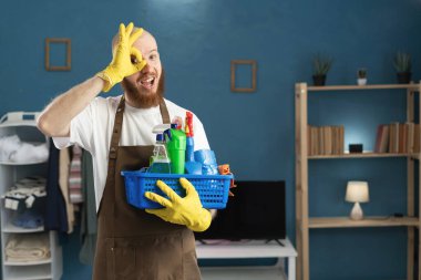 Eldiven ve önlük giyen gülümseyen adam temizlik malzemeleriyle dolu sepet tamam işareti gösteriyor, kameraya katılıyor ve gülüyor. Ev işlerinde erkek hizmetçi. Boşluğu kopyala
