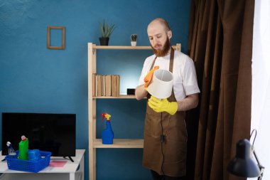 Evde masa lambasının tozunu temizleyen önlük giyen bir adam ev bakımı, günlük ev işi, yüzey hijyeni ve sorumlu oda temizliği kavramını gösteriyor.
