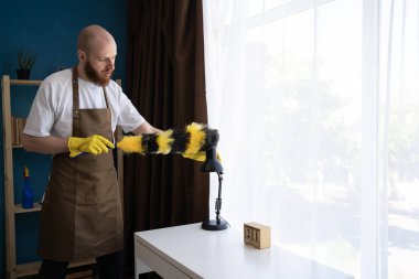 Oturma odasındaki lamba tozunu silmek için tüy tozu kullanan bir adam ev işleri, günlük ev işleri, temizlik rutini ve rahat bir ortamda ev temizliği kavramını gösteriyor.