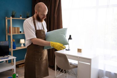 Ev işleri, ev işleri ve temizlik hizmeti konsepti. Önlük takan adam kanepe yastıklarını fırçayla temizliyor, ev temizliği yapıyor. Boşluğu kopyala