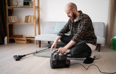 Elinde elektrikli süpürge olan sakallı bir adam ev temizliği, ev temizliği, ev bakımı, hazırlık ve ev işlerinin sorumluluğunu göstermeye hazırlanıyor.