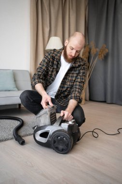 Ev işleri konsepti. Sakallı adam evde elektrik süpürgesinin toz filtresini açıyor. Evi temizledikten sonra. Boşluğu kopyala