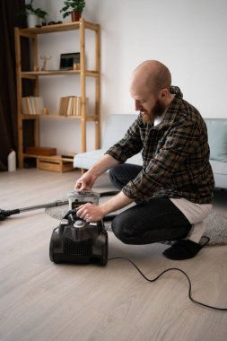 Ev işleri konsepti. Sakallı adam evde elektrik süpürgesinin toz filtresini açıyor. Evi temizledikten sonra. Boşluğu kopyala