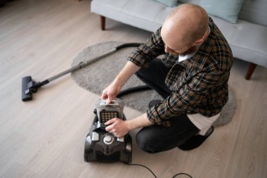 Adam elektrik süpürgesini temizleyince söküyor. Oturma odasında kirli konteynırı boşaltıyor. Ev temizliği kavramı