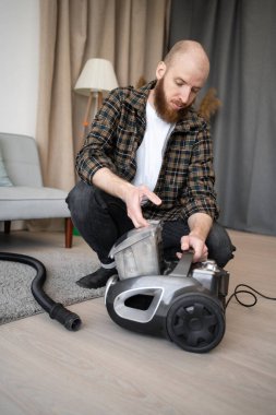 Adam elektrik süpürgesini temizleyince söküyor. Oturma odasında kirli konteynırı boşaltıyor. Boşluğu kopyala