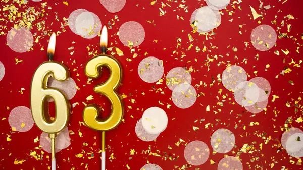 Altmış üç numaralı altın yanan mum. Kırmızı arkaplanda 63 yazılı doğum günü tebrik kartı konfeti ve simle. Yıldönümü, kutlama etkinliği ve doğum günü konsepti.