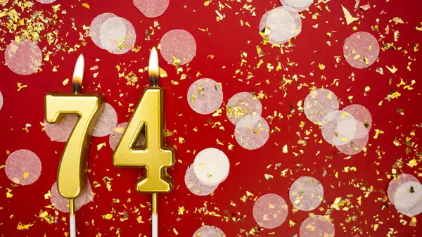 Altın yanan mum numarası 74 'tü. Kırmızı arkaplanda 74 yazılı doğum günü tebrik kartı konfeti ve simle. Yıldönümü, kutlama etkinliği ve doğum günü konsepti.