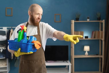 Önlüklü sakallı bir adam, evi temizledikten sonra elinde deterjan sepetiyle seni işaret ediyor. Boşluğu kopyala