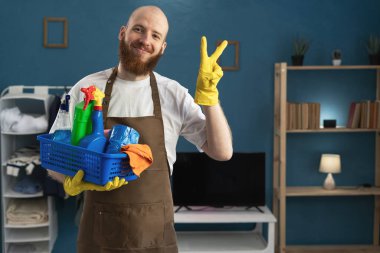 Sakallı hademe zafer jesti yapıyor. Ev işleri için temizlik malzemeleriyle dolu sepeti tutan kameraya bakıyor. Boşluğu kopyala