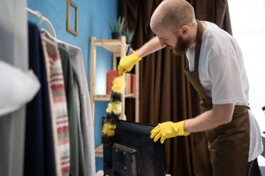 Önlüklü genç sakallı adam, oturma odasında çalışan toz bezi kullanıyor. Boşluğu kopyala