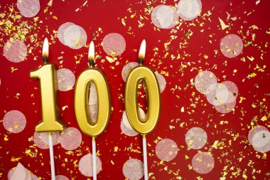 Altın yanan mum 100 numara. Kırmızı arkaplanda 100 yazılı doğum günü tebrik kartı konfeti ve simle. Yıldönümü, kutlama etkinliği ve doğum günü konsepti.