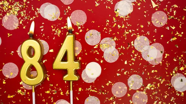 Seksen dört numaralı altın yanan mum. Kırmızı arka planda 84 yazısıyla birlikte konfeti ve simle birlikte doğum günü tebrik kartı. Yıldönümü, kutlama etkinliği ve doğum günü konsepti.