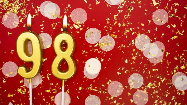 Altın yanan mum numarası 98 'di. Kırmızı arkaplanda 98 yazılı doğum günü tebrik kartı konfeti ve simle. Yıldönümü, kutlama etkinliği ve doğum günü konsepti.