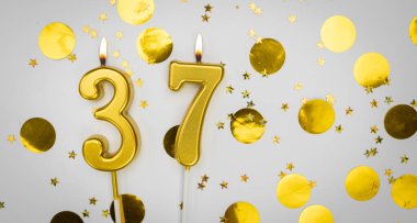 Beyaz arka planda, altın konfeti ve simle birlikte 37 numaralı yanan doğum günü mumu. Otuz yedi yaşında. Doğum günün kutlu olsun. Geçmiş şenliği kartı. Bayrak