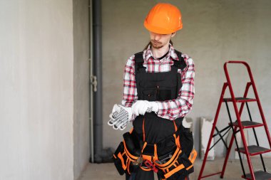 Koruyucu bir miğfer takmış, arka planda iş eldivenleri takan genç bir müteahhit. İnşaat ve tadilat konsepti. Boşluğu kopyala