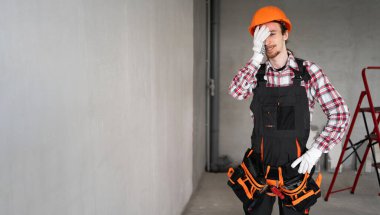 Güvenlik kaskı takmış canı sıkkın bir müteahhit inşaat alanında mutlu bir şekilde acı çekerken yüzünü tutuyor. Boşluğu kopyala