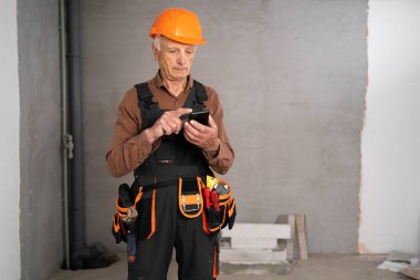 Tamir, inşaat ve inşaat kavramı. Gri arka planda akıllı telefonu olan yaşlı bir inşaatçı. Boşluğu kopyala