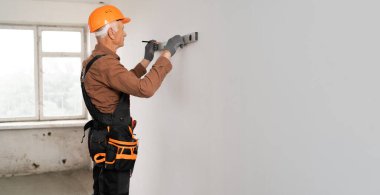 Yaşlı tamirci. Üniformalı kıdemli müteahhit ruh seviyesini kullanarak duvarı ölçüyor ve duvarda izler bırakıyor. Boşluğu kopyala