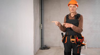 İnşaat alanındaki kıdemli inşaatçı güvenlik kaskı takmış, parmakları fotokopi alanı reklamını ya da terfisini işaret eden el hareketleriyle ayakta duruyor. Yaşlı inşaat işçisi. Pankart. Boşluğu kopyala