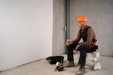 Yaşlı müteahhit ya da tamirci, kafasında kasketle kahve içerek kapalı alanda inşaat alanında oturuyor. Boşluğu kopyala