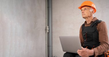 İnşaat alanında dizüstü bilgisayar kullanarak oturan miğferli yaşlı bir inşaatçı. İnşaat, yenileme ve küçük işletme konsepti. Pankart. Boşluğu kopyala