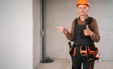 İnşaat alanındaki kıdemli müteahhit güvenlik kaskı takmış, başparmağıyla reklam alanını işaret ediyor. Yaşlı inşaat işçisi. Pankart. Boşluğu kopyala